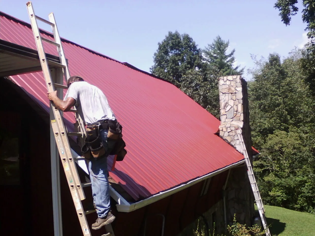 Roofer on a ladder beside a red metal roof and stone chimney — Soffit & Fascia Repair by Roofing Brothers in Blounts Creek